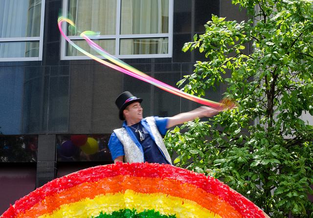 A+man+standing+behind+a+rainbow+parade+float+waves+colorful+banners+at+Boston+Pride+2012.