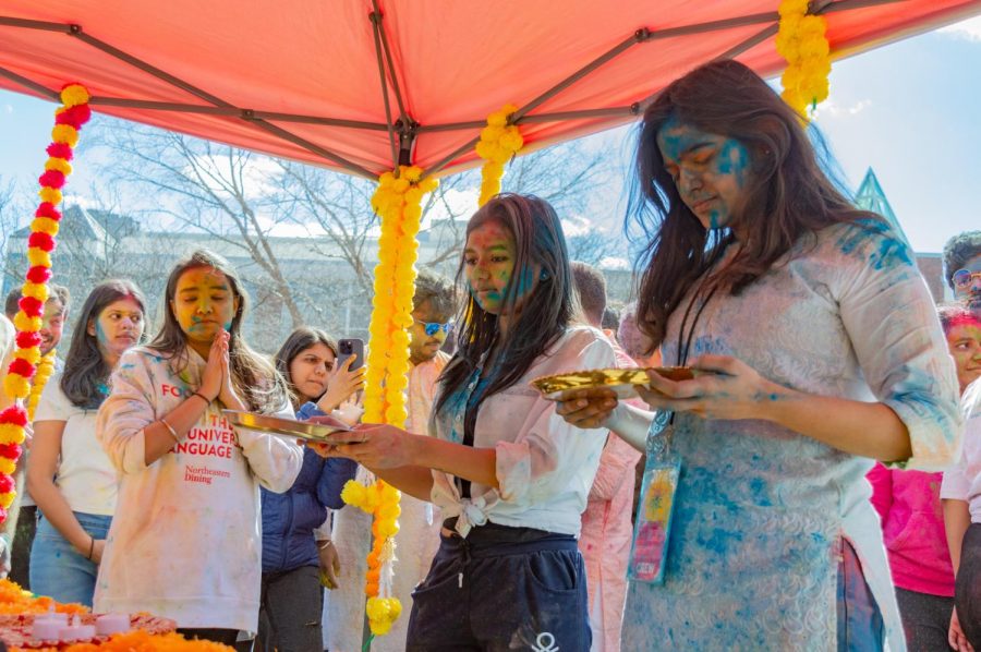 Two students stand under a tent with golden plates to perform the traditional Holi puja.