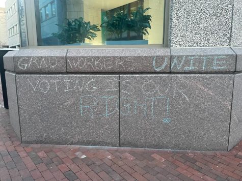 A wall on campus with chalk messaging promoting graduate students right to unionize. The union representing grad students has filed charges against Northeastern after university officials allegedly illegally retaliated against students for chalking.