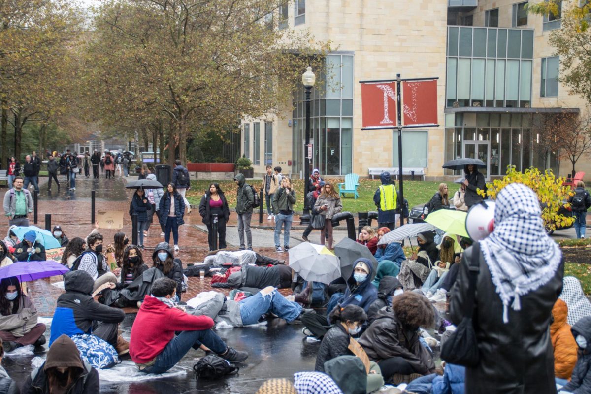 An activist addresses students and faculty passing by the die-in via megaphone, inviting them to join the protest. Several passersby stopped to watch or film the event, or to talk with activists.