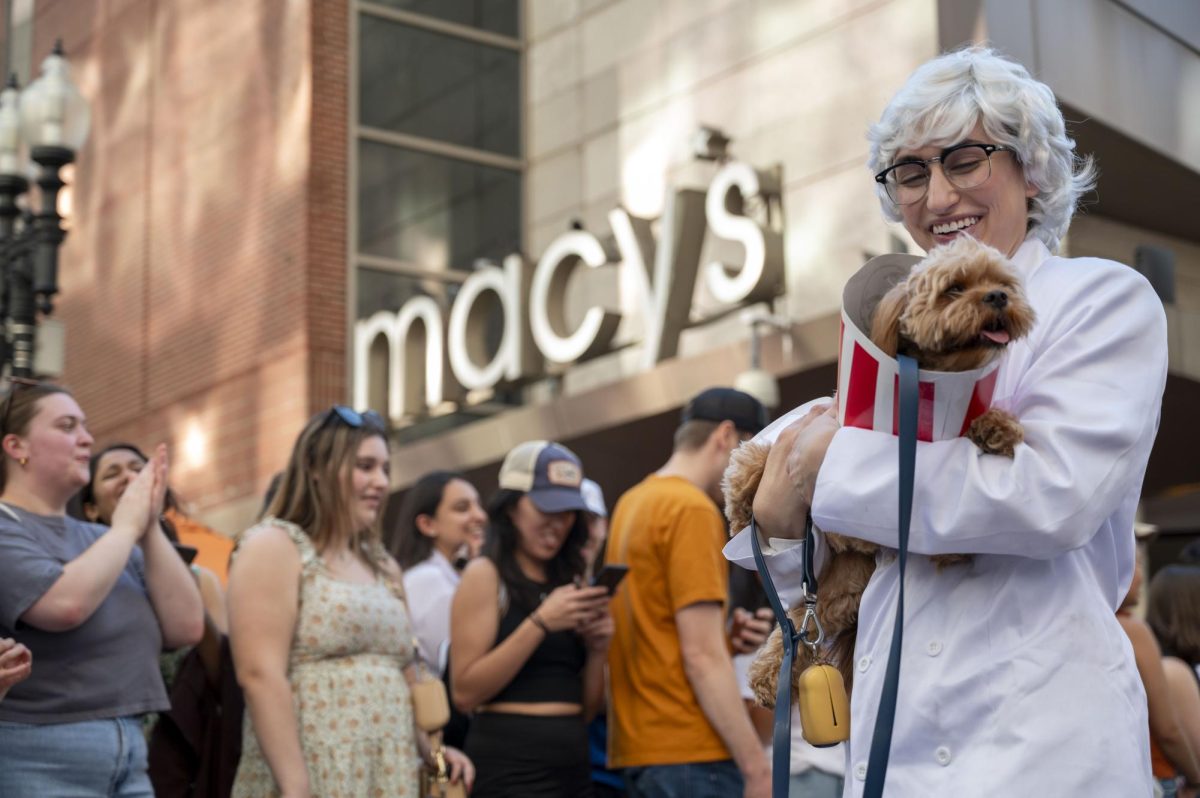 A dog owner dressed as Colonel Sanders holds his dog, dressed as a bucket of fried chicken, while they walk past the crowd. Some dogs were carried in the parade while others were pushed in a stroller.