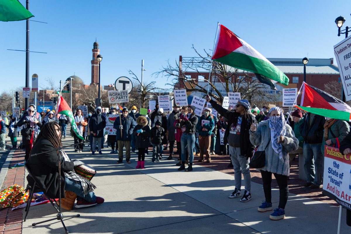 A crowd with Palestinian flags and signs reading End all U.S. aid to Israel and Stop bombing Palestine, Yemen, Syria and Lebanon gather around a man playing a Ghanian drum. Hundreds gathered at Roxbury Crossing Sunday to protest Northeastern University and other colleges role in the Israel-Palestine war.