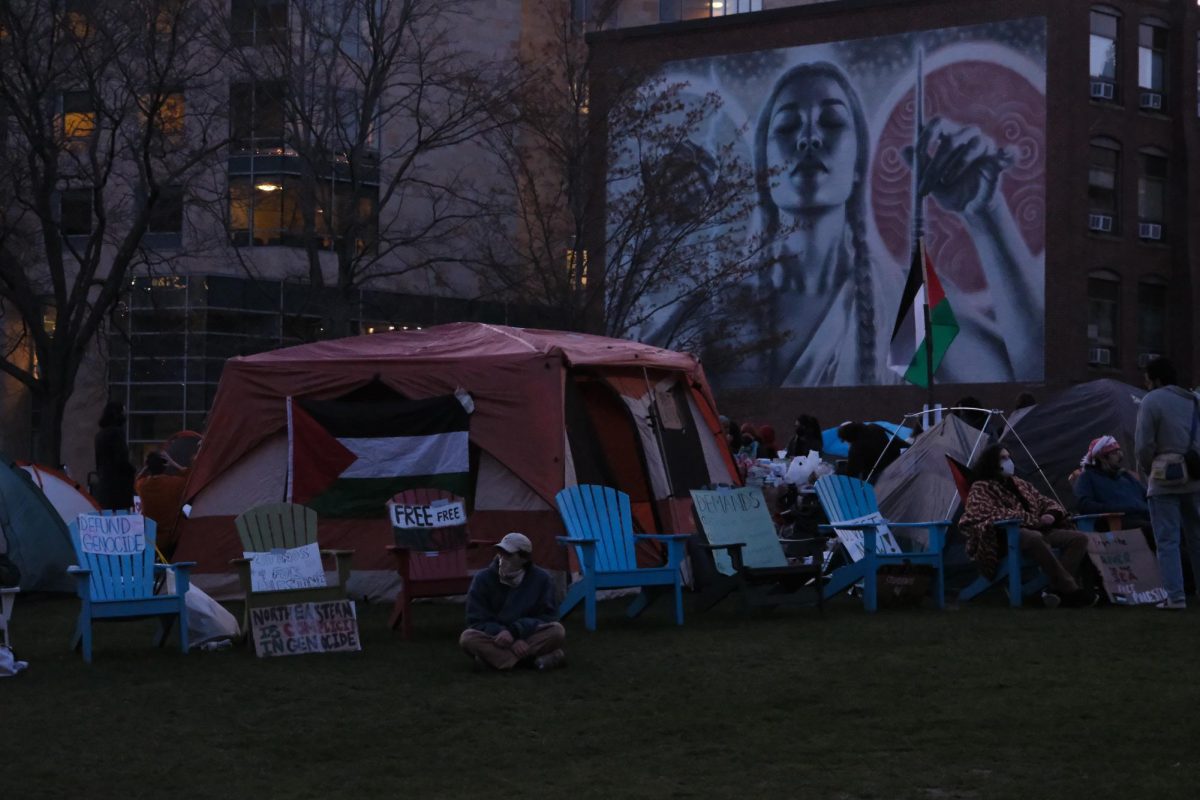 A protester surrounded by signs and Adirondack chairs sits in the encampment. Renata Nyul stated that the power was shut off out of safety concerns for demonstrators.