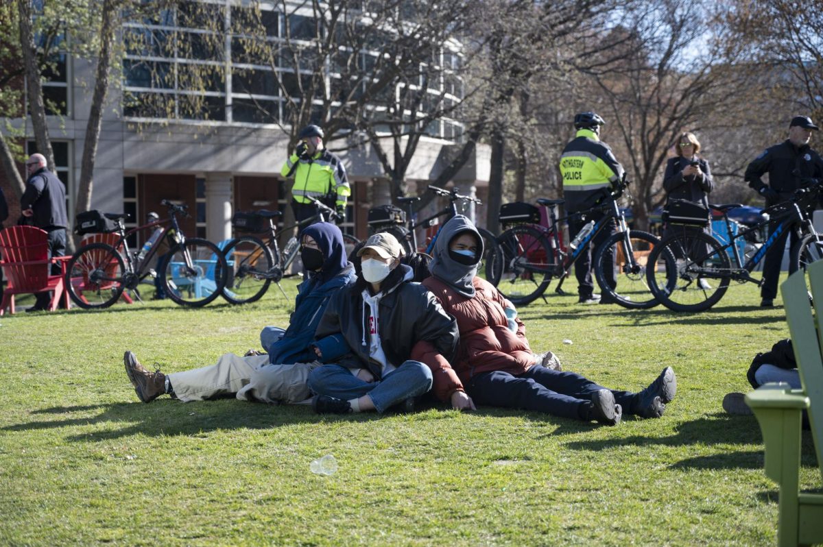 Three students link arms and chant pro-Palestine slogans on Centennial Common while surrounded by police officers and a circle of chairs and bikes April 25. Following the start of the demonstration around 8 a.m., Huskies for a Free Palestine, or HFP, called for additional students to join protesters on Centennial.