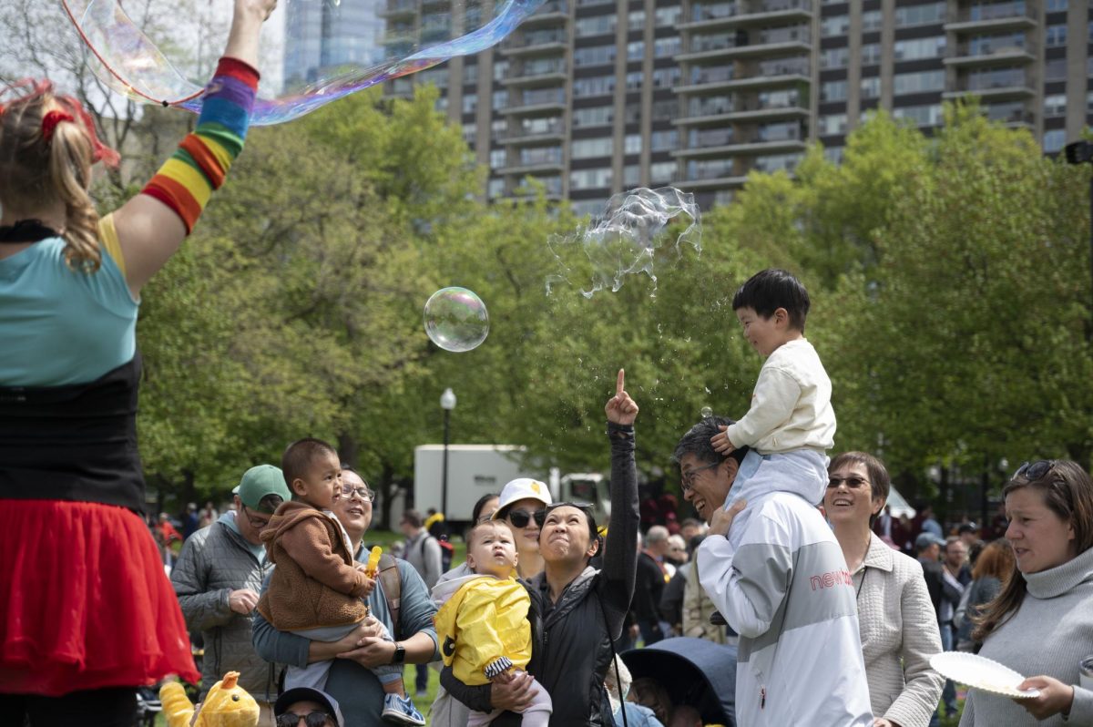 A mother pops a bubble with her finger as children and parents around her watch and smile. Jenny the Juggler stood atop a tall platform and continuously threw large bubbles by dipping a long rope in bubble solution.