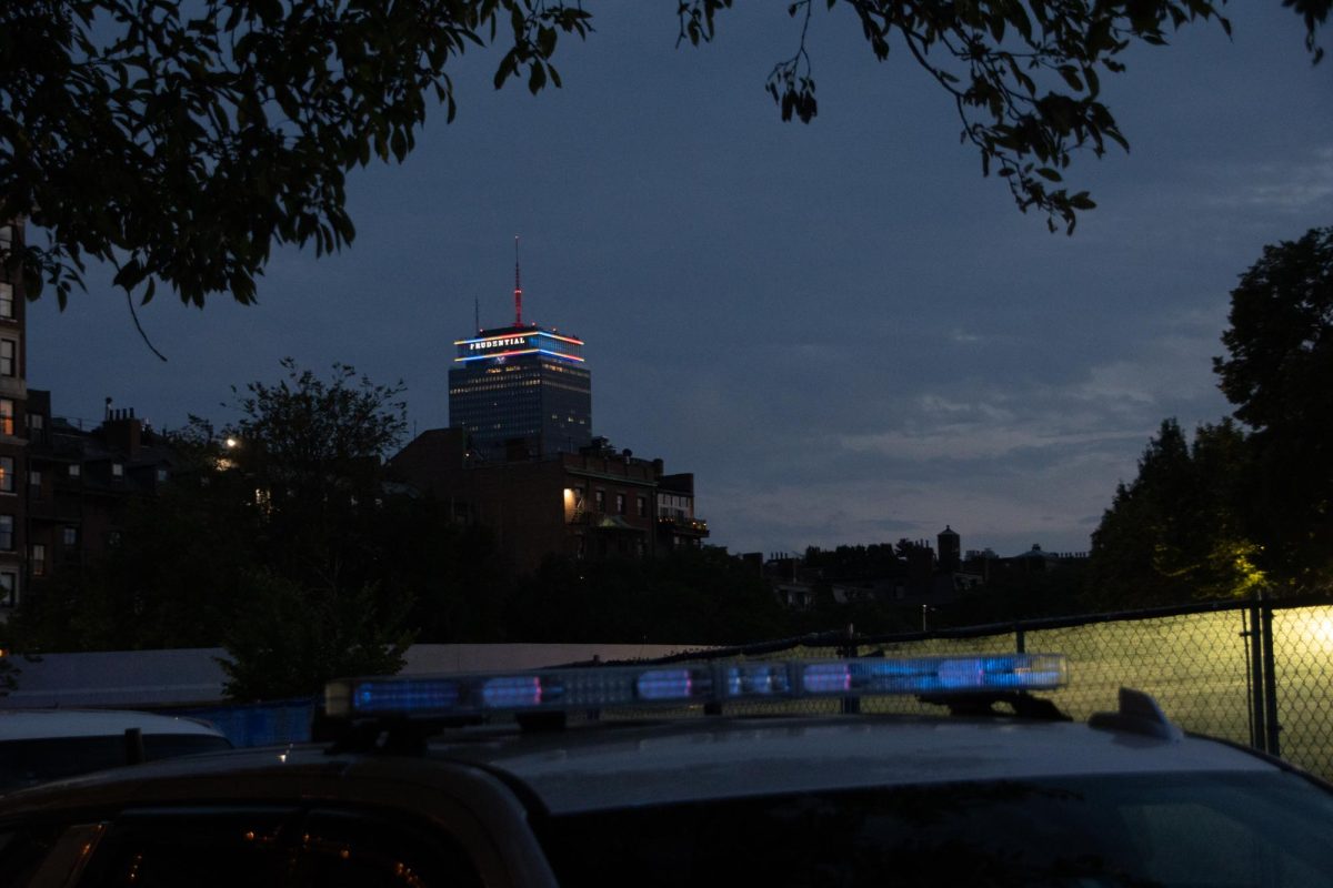 The Prudential Tower glows in the colors of the United States flag, as seen across the top of a parked police car and a chain-link fence. As one of the cities where the first battles of the Revolutionary War occurred, Boston takes every Fourth of July as a chance to show off its deep sense of patriotism.