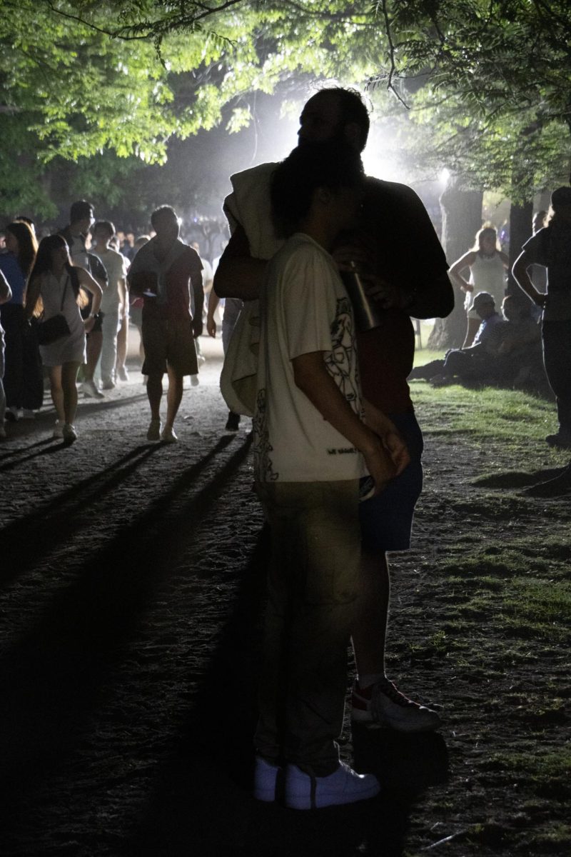 The harsh glow from a set of portable spotlights throws shadows across a group of spectators waiting in line for the bathroom. Boston authorities brought in external lighting to illuminate the Charles River Bank for safety, as the popular spot for joggers and tourists gets dark at night.