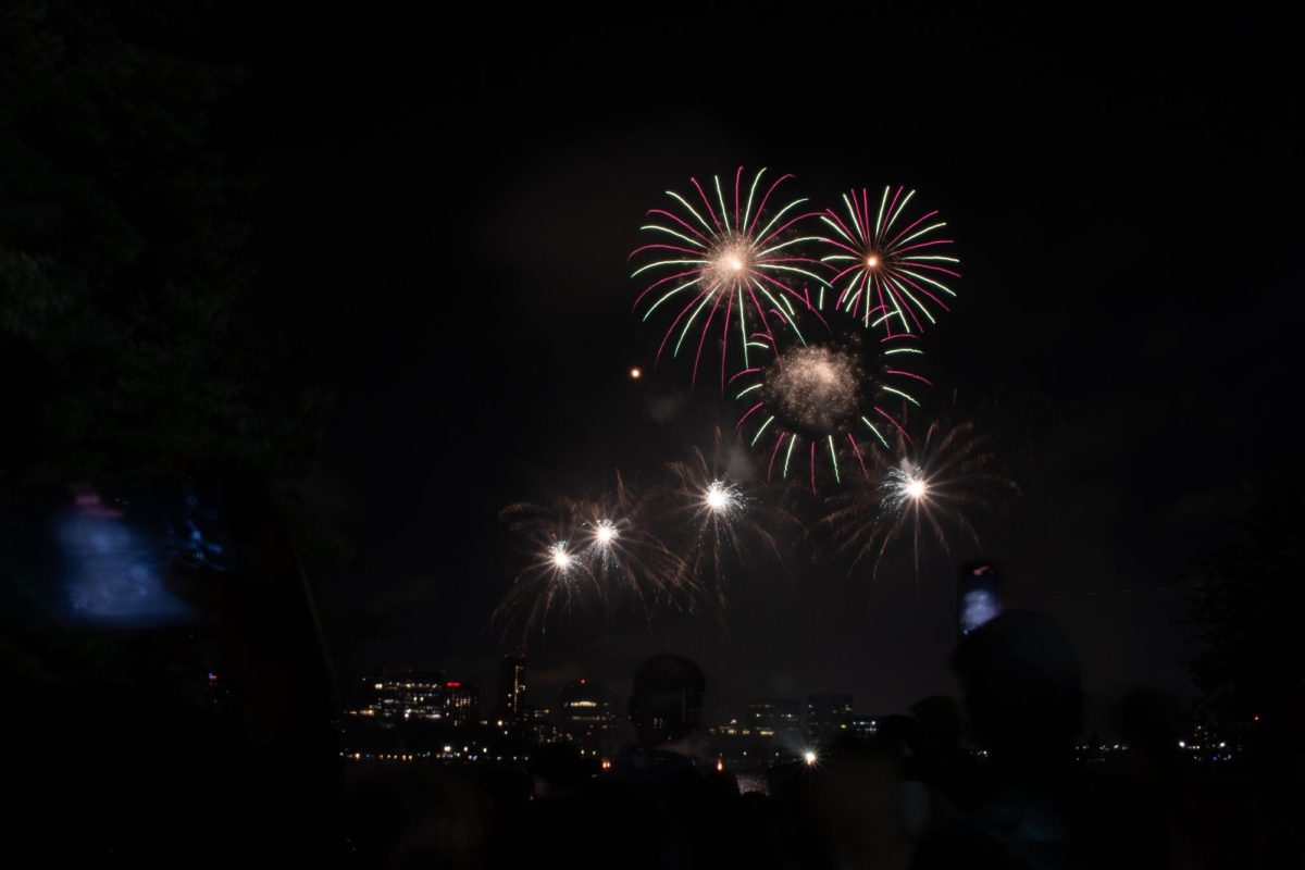 Pale green streaks of light mingle with red in a circular fireworks burst, with lights of the skyline visible below. Spectators crowded onto fire escapes, balconies and roofs to catch a glimpse of the Fireworks Spectacular.