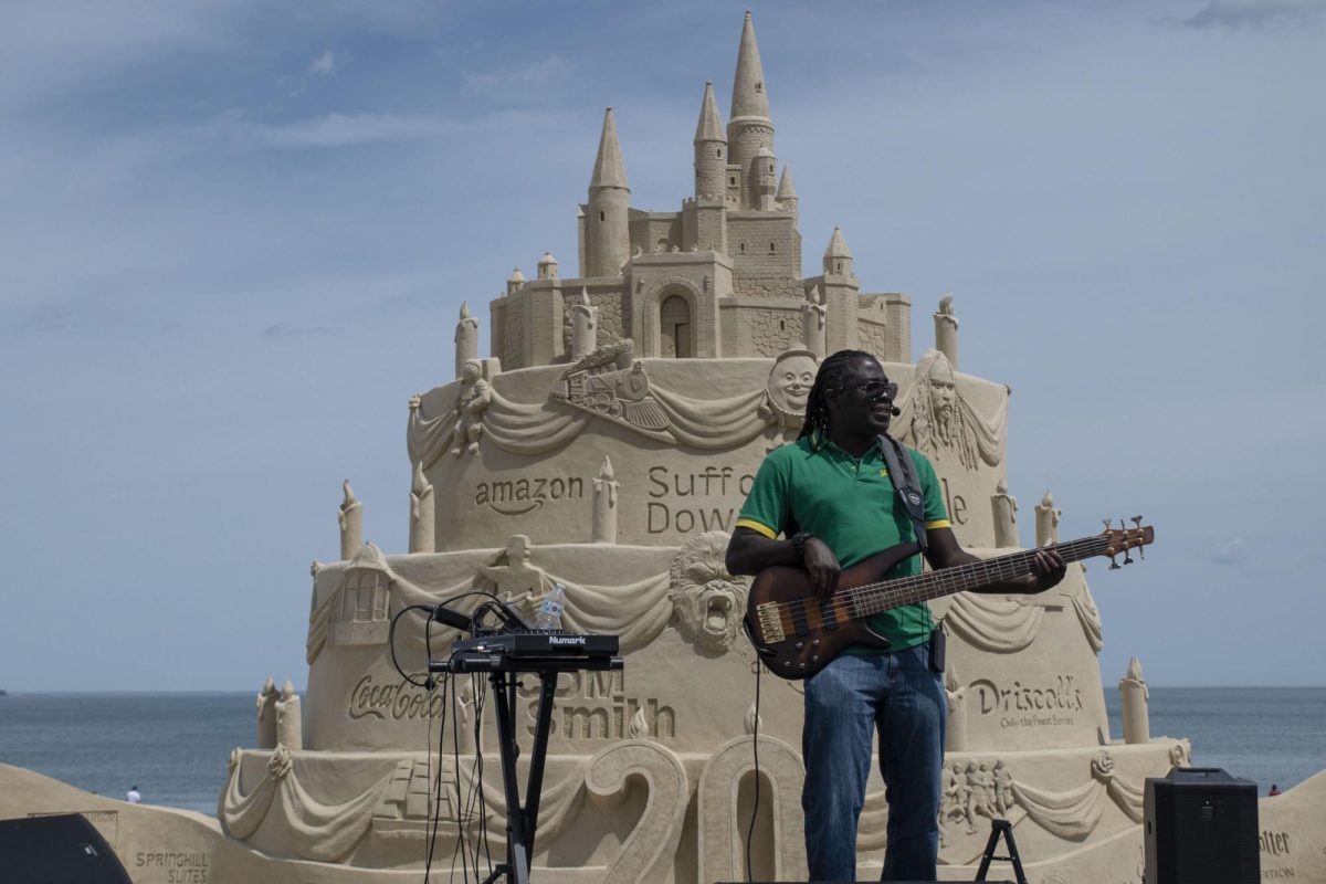 A performer sings and plays the bass guitar in front of a 20th anniversary cake sand sculpture with a sand castle on top. The cake sculpture was the centerpiece of the event and contained tributes to past festival centerpieces.