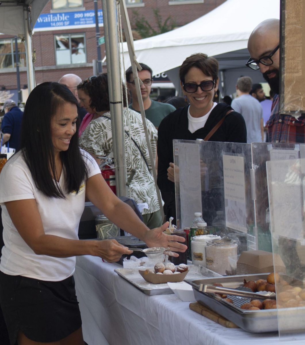 A vendor sprinkles powdered sugar over a tray of fried dough balls while customers watch. Over 80 vendors attended the Boston Local Food Festival.