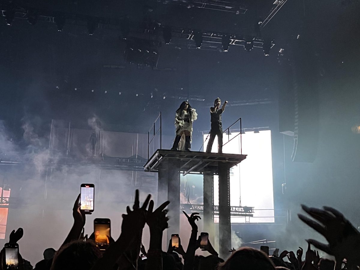 Charli XCX and Troye Sivan stand on a raised platform as fans cheer and raise their hands. The two performed at TD Garden Sept. 28 for their "Sweat Tour."