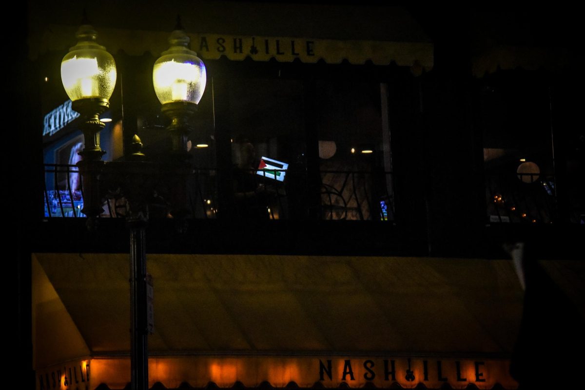 An individual in a nearby restaurant holds up a Palestinian flag on their electronic device in support of the picket. Bystanders watched the picket as they passed by on sidewalks or from vantage points inside nearby buildings like the Nash Bar & Stage.