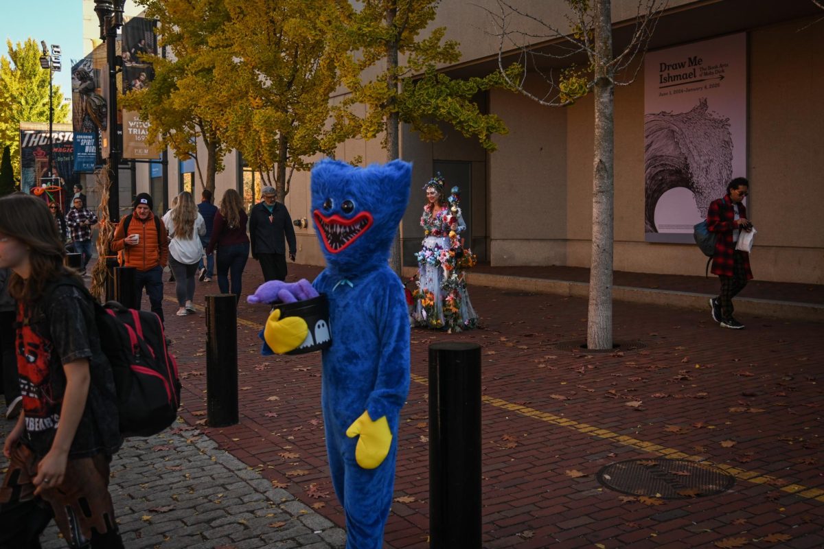 Individuals, including a costumed Huggy Wuggy monster from the popular game Poppy Playtime passing in front of a costumed fairy-like figure, make their way through the marketplace. People of all ages, from performers to those dressing up for fun, walked through the densely-packed area.