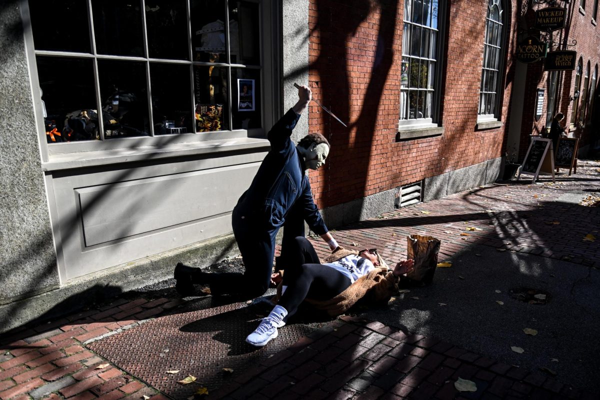 A person costumed as Michael Myers playfully pretends to stab a woman posing for a picture outside a building on Essex Street. Performers and individuals dressed up in costumes, including notable fictional horror characters like Myers, were scattered throughout the marketplace and would switch spots as the day progressed, leaving bags and buckets for tips after posing for pictures.