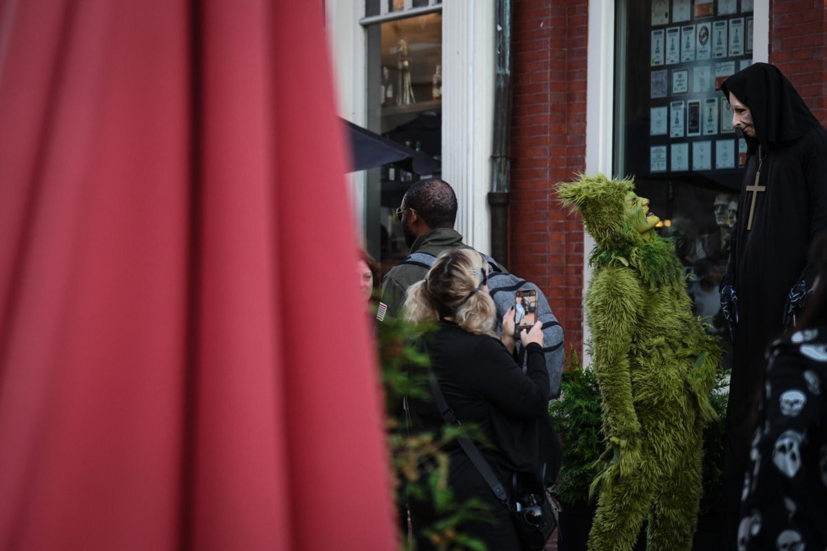 A pedestrian holds up their phone to capture an individual dressed as the Grinch interacting with a taller costumed figure in the marketplace. Those in costumes not only interacted with passersby, but also with each other, drawing further attention from onlookers.