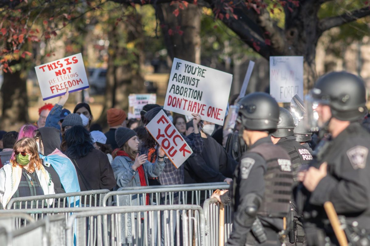 Counterprotesters wave signs from behind a row of metal barricades as BPD officers in riot gear look on. Pro-choice activists carried dozens of handmade signs, including some in the shape of clothing hangers, with slogans that railed against religious dogma, anti-abortion activism and perceived government control of their bodies.