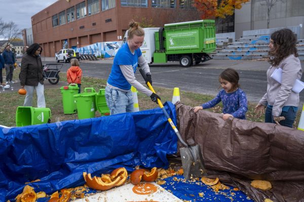 A Mothers Out Front volunteer shovels smashed pumpkin pieces as a child participant smiles and watches. Volunteers picked up larger pumpkin pieces and gave them to children to smash into the pit again.