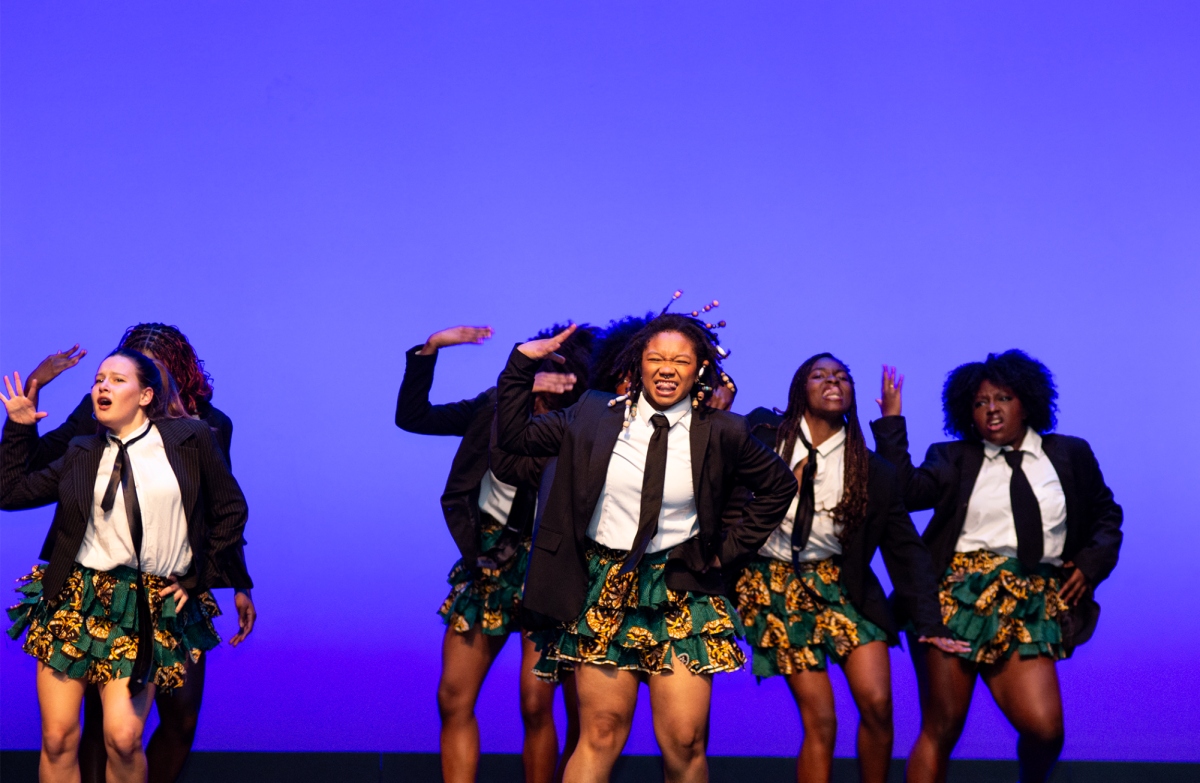 The Omo Naija x Wahala Boys perform in matching outfits — black blazers, ties, white shirts and patterned ruffled skirts — in front of a blue background. This team was the fifth to compete.