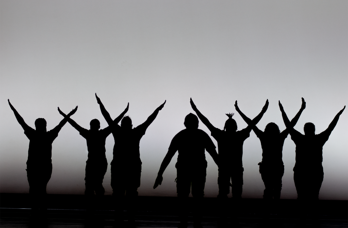 Silhouettes of the SOAL Step Team stand in formation creating “Y” shapes in front of a gray background. The team opened the show with their incredible and fierce performance.