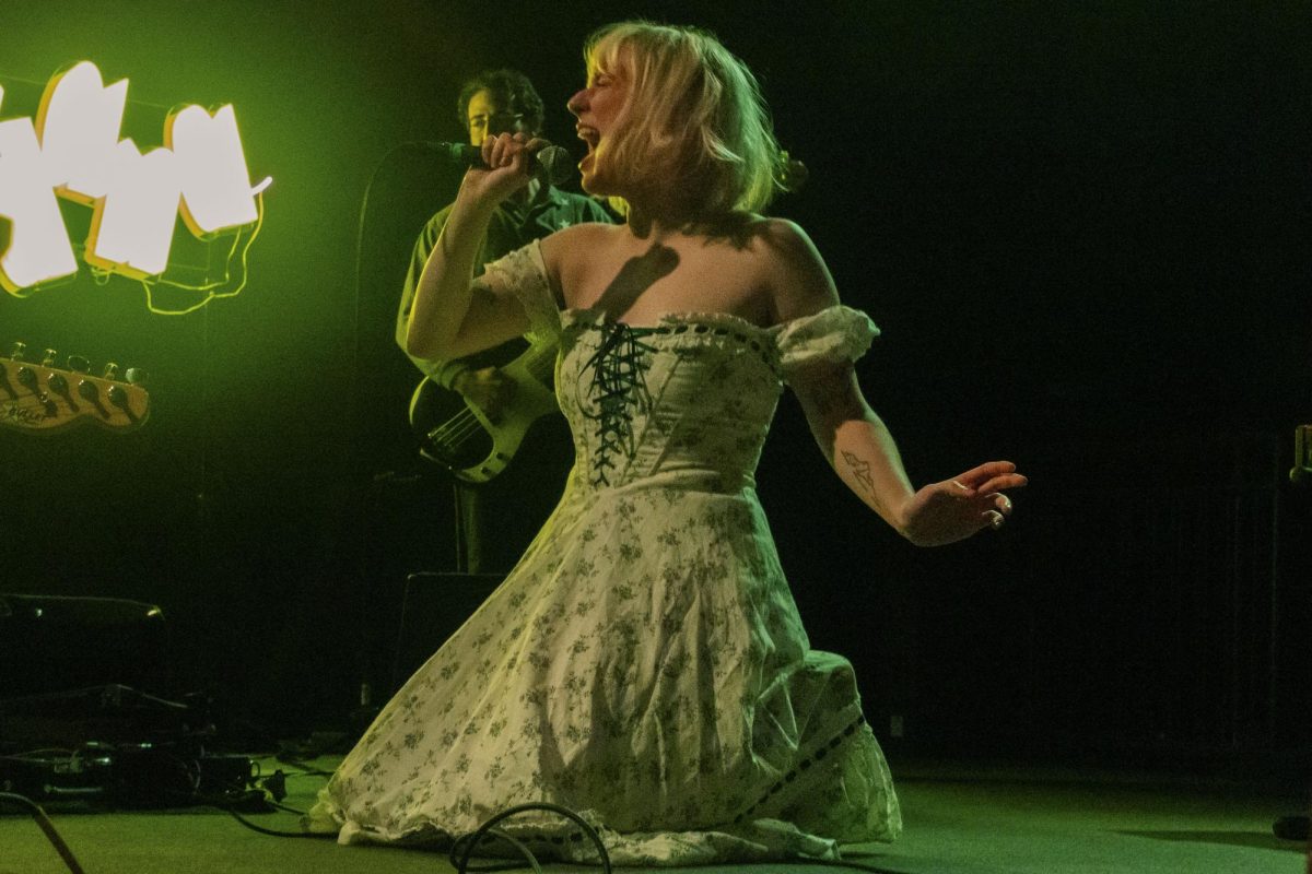 Jordana sings while kneeling on the ground against a neon green light. She played the violin, guitar and tambourine during the show.