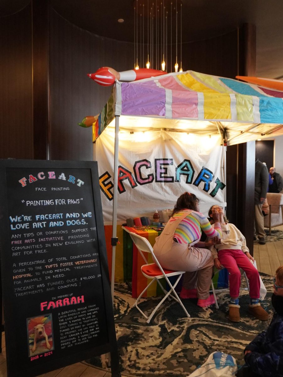 A face painter begins to paint on a small child’s cheek. A long line formed in front of the stand with many children waiting to participate.